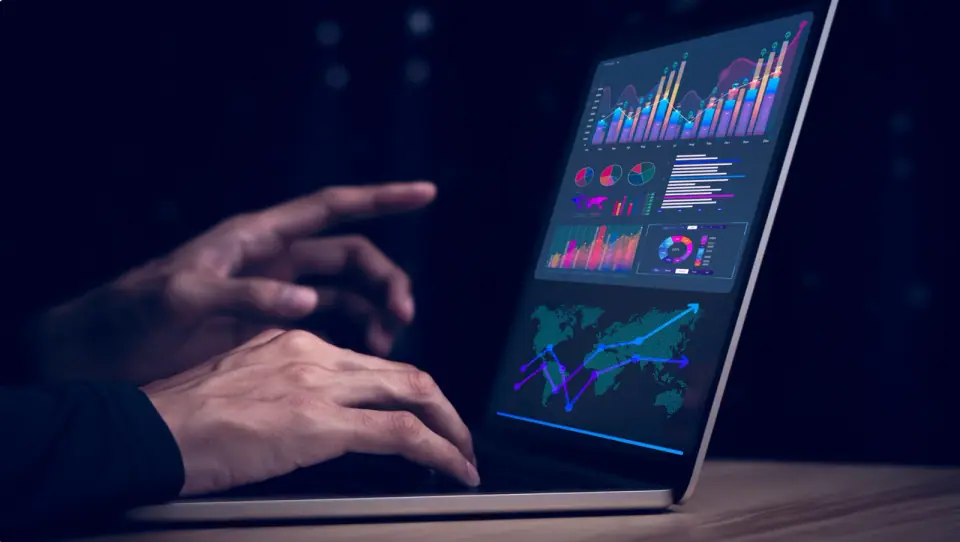 Hands at a laptop in a dark room, the screen filled with operations dashboards: bar and line charts, pie charts and a world map of trend lines